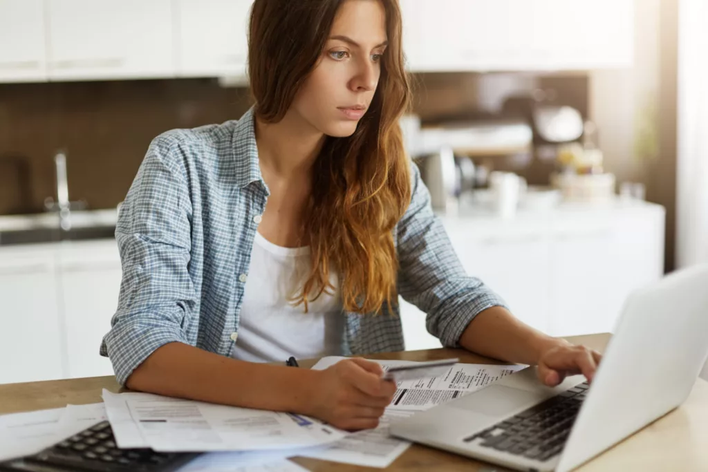 Pretty woman with long loose hair surfing websites while shopping online, using credit card to pay for online purchase, looking at laptop screen with concentrated expression. Modern technologies